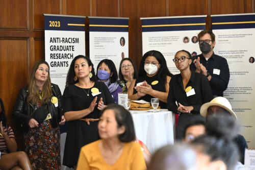 Students and staff listen and applaud a speaker as they stand in front of anti-racism research posters